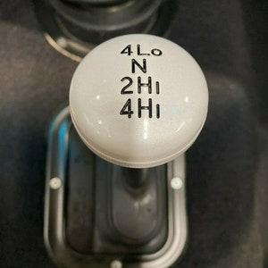 May include: White gear shift knob with black lettering indicating the different gear positions: 4LO, N, 2HI, 4HI.