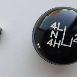 May include: A black transfer case shift knob with white lettering indicating 4L, N, 4H, and 2H positions. A silver hex nut is to the left of the knob. The objects are on a white surface.