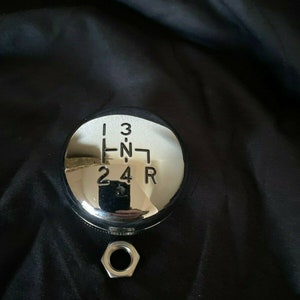 May include: Chrome gear shift knob with a black background. The knob has a circular design with the numbers 1, 2, 3, 4, R, and N engraved on it.