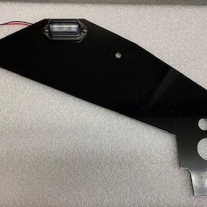 May include: Black metal bracket with a small clear light and wiring. The bracket has several holes for mounting.