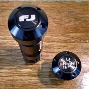 以下が含まれることがあります： 銀色のFJロゴとH、N、L、4の文字が書かれた黒いシフトパターンが付いた黒いシフトノブ