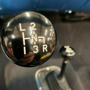 May include: Black gear shift knob with white lettering for a car. The lettering reads "L 2 -N- 1 3 R".