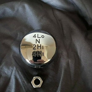 May include: A silver gear shift knob with the text "4Lo, N, 2Hi, 4Hi" on it. A silver nut is also in the image.
