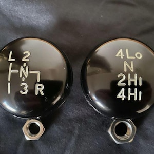 May include: Two black round gear shift knobs with silver lettering. The left knob has the letters L, 1, 2, 3, R, and N. The right knob has the letters 4Lo, N, 2Hi, and 4Hi.