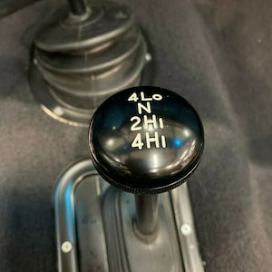 May include: A black gear shift lever with a round knob that has white lettering indicating the different gear positions: 4LO, N, 2HI, 4HI.