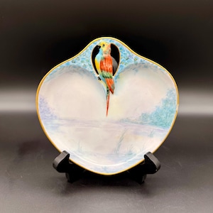 May include: A decorative, oval-shaped ceramic dish with a colorful parrot perched in the center. The dish features a light blue and white landscape design with a gold rim. The parrot is painted in vibrant shades of yellow, blue, and red.