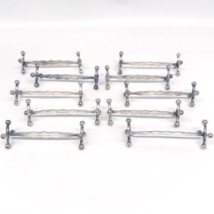 May include: A set of eight silver-toned metal knife rests, each featuring a long, scalloped central bar supported by four small spherical feet at the ends. The metallic surface has a polished finish with visible hallmarks stamped on the center.