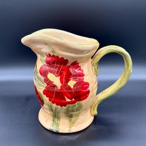 May include: A ceramic pitcher with a beige base and a green handle. The pitcher is decorated with large red floral designs with yellow centers and green leaves. The pitcher has a spout for pouring.