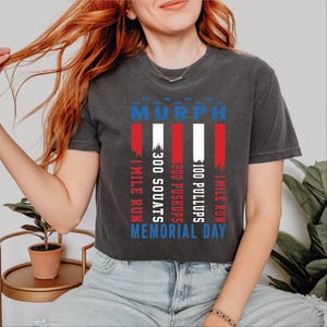 May include: Dark gray t-shirt with a patriotic design. The shirt features the word "MURPH" in blue, with red and white stripes and the text "1 MILE RUN, 300 SQUATS, 200 PUSHUPS, 100 PULLUPS, 1 MILE RUN, MEMORIAL DAY" in white.