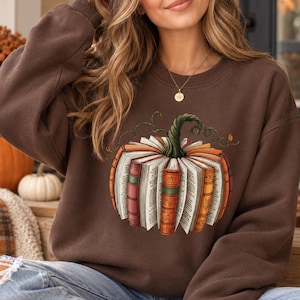 May include: A dark brown crewneck sweatshirt featuring a graphic print of a pumpkin constructed from open books with colorful spines and a green stem. The sweatshirt has long sleeves and a ribbed collar, cuffs, and hem, providing a casual fit.