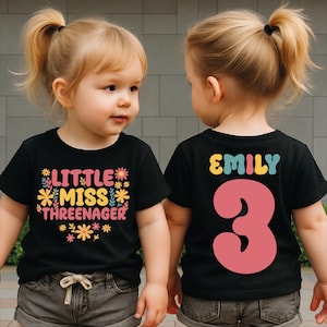 May include: Two toddlers wearing black t-shirts. The front of one shirt says "LITTLE MISS THREENAGER" with floral accents. The back of the other shirt says "EMILY 3" in colorful letters and a large pink number. The girls are wearing gray shorts.