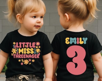 May include: Two toddlers wearing black t-shirts. The front of one shirt says "LITTLE MISS THREENAGER" with floral accents. The back of the other shirt says "EMILY 3" in colorful letters and a large pink number. The girls are wearing gray shorts.