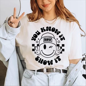 May include: White t-shirt with a black and white graphic design. The graphic features a smiling face wearing a hat with the word "NOW" on it. The text "YOU KNOW IT SHOW IT" is written in a retro font around the face.