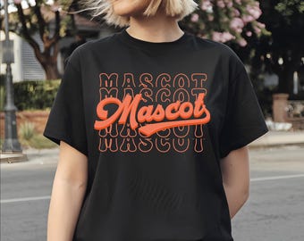 May include: Black t-shirt with the word "Mascot" in orange, stacked vertically with a retro font. The word "Mascot" is also written in a larger, cursive font in the center of the design. The shirt is a crew neck and has short sleeves.