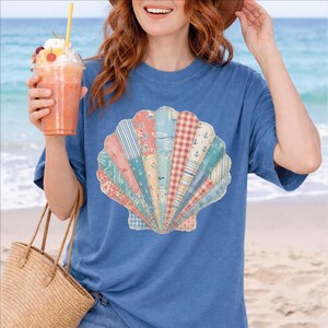 May include: A blue t-shirt featuring a seashell design. The seashell is composed of various patterned fabrics in shades of pink, blue, and white. The image evokes a beach theme, with a person holding a drink and a straw bag.
