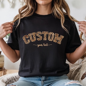 May include: Black t-shirt with the word "CUSTOM" in a leopard print design. Below the word "CUSTOM" is the text "your text" in a cursive font. The t-shirt is a short-sleeved crew neck.
