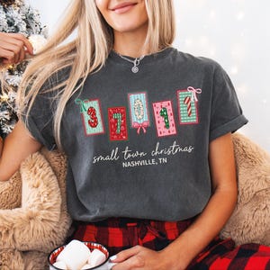 May include: Dark gray t-shirt with a Christmas-themed design. The shirt features colorful patches with numbers and candy canes, along with the text "small town christmas" and "Nashville, TN".