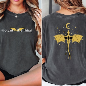 May include: A dark gray t-shirt with the text "violent little thing" on the front and a yellow silhouette of a dragon with wings spread and a crescent moon above it on the back.