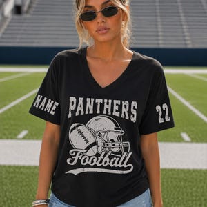 May include: A black V-neck t-shirt featuring a white distressed graphic of a football helmet and a football. The word PANTHERS is printed above the graphic, with the word Football below. The right sleeve says NAME and the left sleeve says 22.