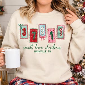 May include: A cream-colored crewneck sweatshirt featuring a graphic of five rectangular patches with festive patterns and numbers. Below the patches, the text reads "small town christmas" and "NASHVILLE, TN" in green lettering.