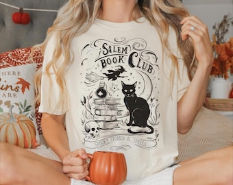 May include: Cream-colored t-shirt featuring a black graphic for the Salem Book Club. The design includes a black cat, books, a witch on a broom, and the words "Spooky Stories & Spells." The person is holding an orange pumpkin-shaped mug.