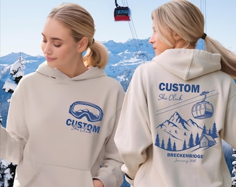 May include: Beige hoodie with a blue ski goggles graphic and "Custom Ski Club" text on the front. The back features a mountain scene with a ski lift, cabin, and text that reads "Breckenridge January 2026".