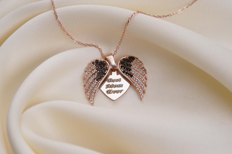 A necklace with a pendant featuring a heart-shaped design with wings and the words "Best Mom Ever" engraved on it, placed on a white fabric background.