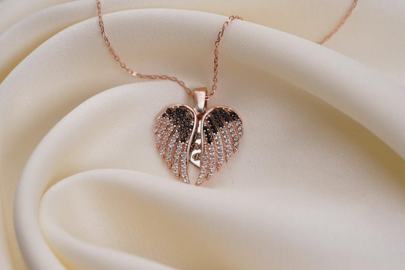 A delicate necklace with a heart-shaped pendant featuring a pair of sparkling wings rests on a soft, white fabric.