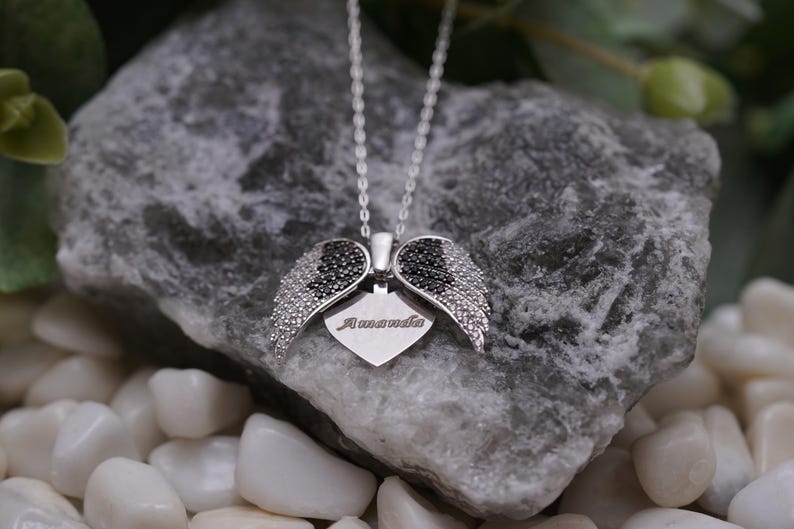 A silver necklace with a heart-shaped pendant featuring black gemstones is displayed on a rock, surrounded by small white pebbles.