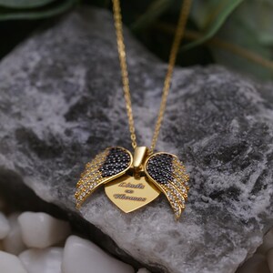A gold-colored necklace with a pendant featuring a pair of black and gold wing-shaped charms, resting on a gray rock.