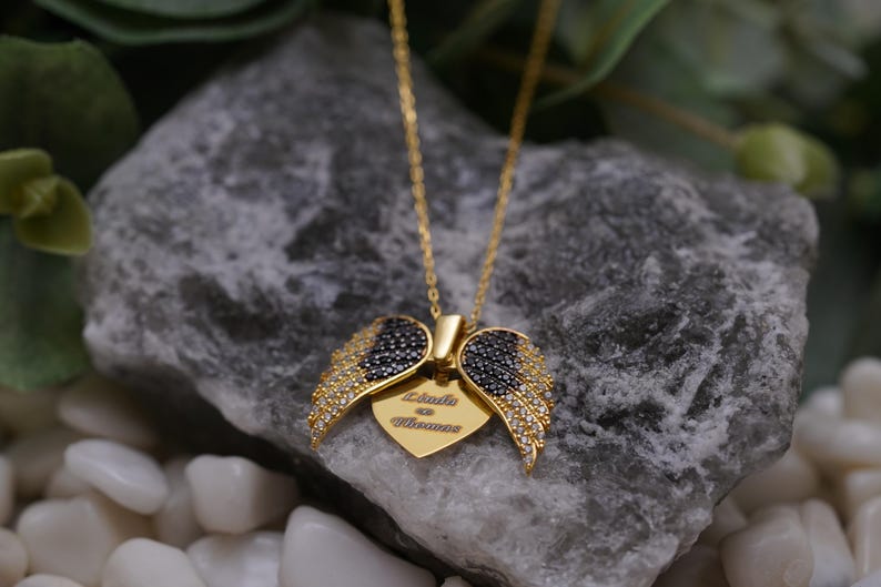 A gold-colored necklace with a pendant featuring a pair of black and gold wing-shaped charms, resting on a gray rock.