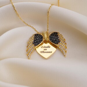A gold-colored necklace with a pendant featuring a heart-shaped design and diamond wings, placed on a white fabric background.