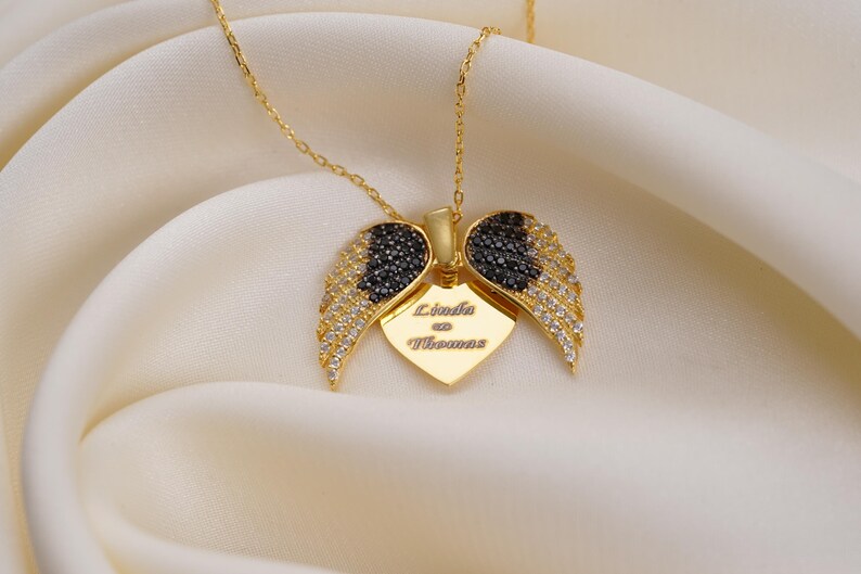 A gold-colored necklace with a pendant featuring a heart-shaped design and diamond wings, placed on a white fabric background.