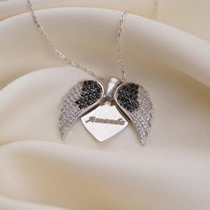 A silver necklace with a pendant featuring a heart-shaped design and diamond wings, placed on a white fabric background.
