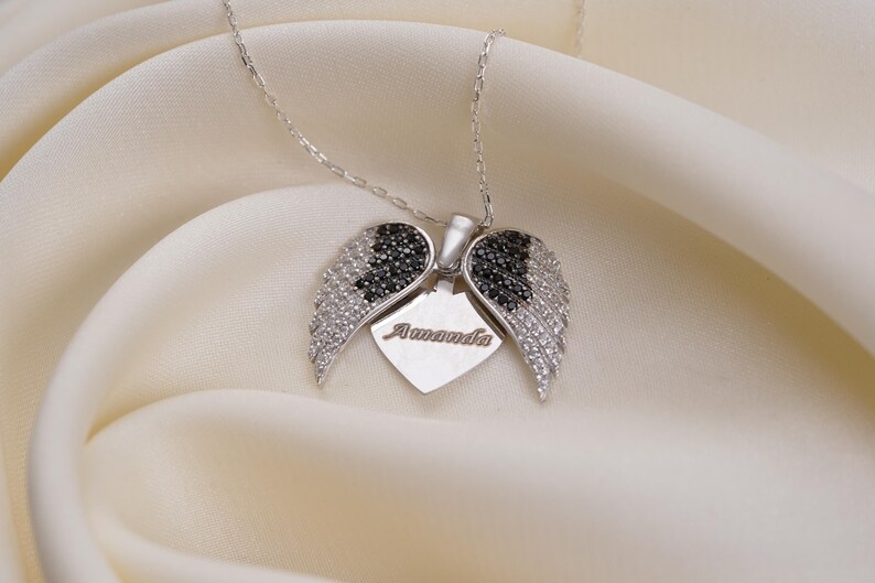A silver necklace with a pendant featuring a heart-shaped design and diamond wings, placed on a white fabric background.
