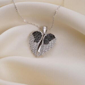 A silver necklace with a heart-shaped pendant adorned with black and white diamonds is displayed on a white fabric surface.