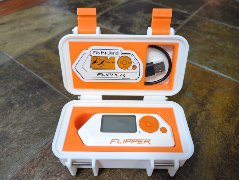 May include: A white and orange case with a small white and orange device inside. The device has a screen, a button, and the word "FLIPPER" printed on it. The case has a USB cable inside and the words "Flip the World!" and "FLIPPER" printed on it.