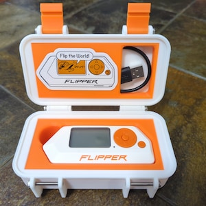 May include: A white and orange case with a small white device inside. The device has a screen, a button, and the word "FLIPPER" printed on it. The case has a USB cable inside and the words "Flip the World!" printed on the inside of the lid.