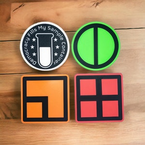 Op de afbeelding: Vier ronde stickers met verschillende ontwerpen. De eerste sticker is zwart met witte tekst die "Democracy Fills My Sample Container" luidt en een wit reageerbuispictogram. De tweede sticker is groen met een zwarte lijn in het midden. De derde sticker is oranje met een zwart vierkant erin. De vierde sticker is rood met vier zwarte vierkanten erin.