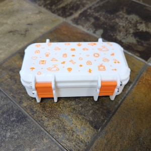 May include: A small white rectangular box with orange latches. The box is decorated with a variety of small orange icons, including a whale, a house, a cloud, a star, a heart, a tree, a bicycle, and a television.