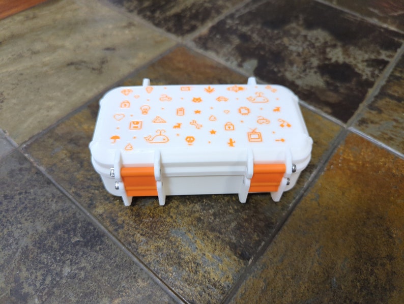 May include: A small white rectangular box with orange latches. The box is decorated with a variety of small orange icons, including a whale, a house, a cloud, a star, a heart, a tree, a bicycle, and a television.
