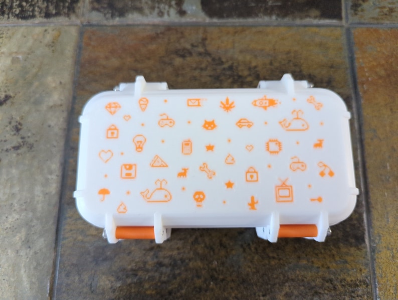 May include: A white rectangular storage container with orange latches. The container has a white background with a pattern of orange icons, including a whale, a lightbulb, a heart, a star, a bone, a cactus, a TV, and a car.