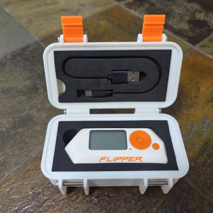 May include: A white and orange case with a small white device inside. The device has a screen and a single orange button. The text "FLIPPER" is printed on the device.