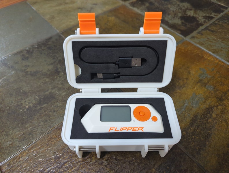 May include: A white and orange case with a small white device inside. The device has a screen and a single orange button. The text "FLIPPER" is printed on the device.