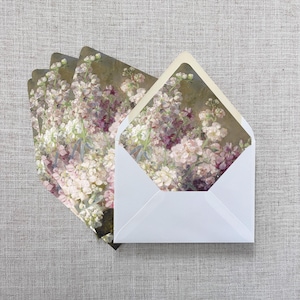 May include: White envelopes with floral print inserts. The inserts feature a painting of white and purple flowers. The envelopes are arranged on a neutral-colored surface, creating a soft and elegant aesthetic. Ideal for stationery and special occasions.
