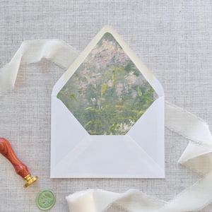 Puede incluir: Un sobre blanco con forro estampado floral, abierto para revelar el diseño interior. Se ve un sello de cera de madera y oro y un sello de cera verde con una mariposa. Una cinta color crema está drapeada alrededor del sobre.