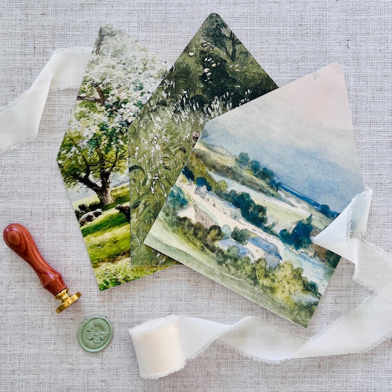 May include: Three landscape-themed greeting cards with watercolor-style artwork. The cards are in a diamond shape, with images of trees, fields, and a rural scene. A wooden handled wax stamp, a green wax seal, and a white ribbon are also present.