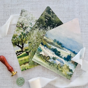 May include: Three watercolor painted envelopes with different landscape scenes. The envelopes are folded and stacked on top of each other. The top envelope features a blue sky with a green field and trees. The middle envelope features a green field with white flowers and trees. The bottom envelope features a green field with a white tree and a blue sky.