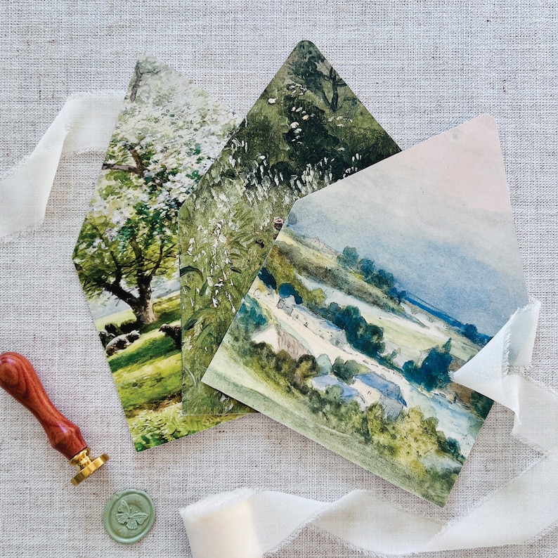 May include: Three watercolor painted envelopes with different landscape scenes. The envelopes are folded and stacked on top of each other. The top envelope features a blue sky with a green field and trees. The middle envelope features a green field with white flowers and trees. The bottom envelope features a green field with a white tree and a blue sky.