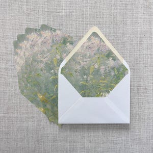 May include: White envelopes with a floral design on the inside, with a matching floral design on the cards. The cards and envelopes are arranged on a gray linen surface. The floral design features shades of green, yellow, and pink.